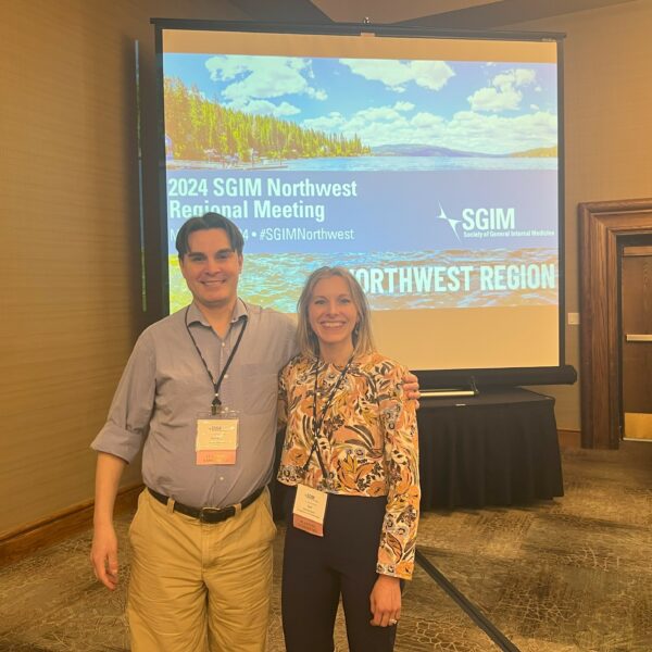 Stefanie Deeds & Patricio Riquelme pose together in front of a projector screen at the 2024 SGIM Northwest Regional Meeting.