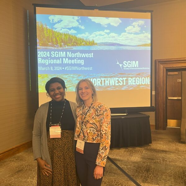 Stefanie Deeds & Ryan Abe pose in front of a projector screen at the 2024 SGIM Northwest Regional Meeting.