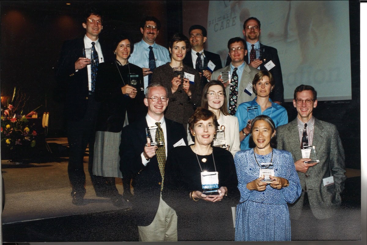Thirteen award recipients in business attire pose in three rows on a stage, each holding an engraved glass award.