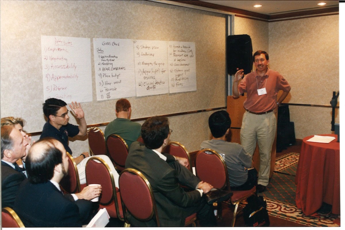 A man presents beside four handwritten flip chart sheets listing attributes and responsibilities of a division chief while seated attendees look on, one with a hand raised.
