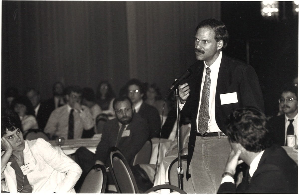 Black and white photo of a man in a suit standing at a floor microphone addressing a seated audience in a banquet hall.