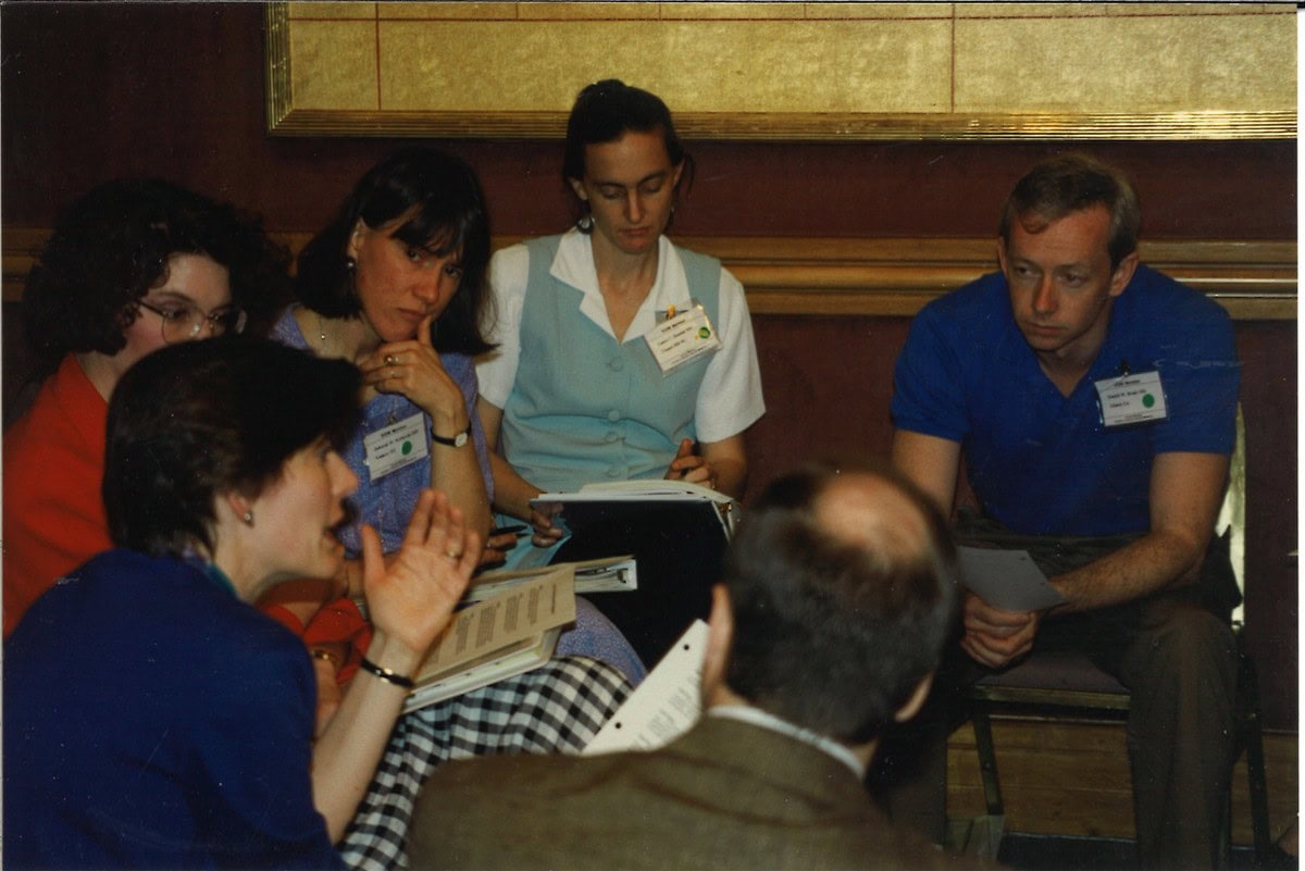 Five people seated in a close circle at a conference session, several holding open binders and handouts while one woman gestures mid-conversation.