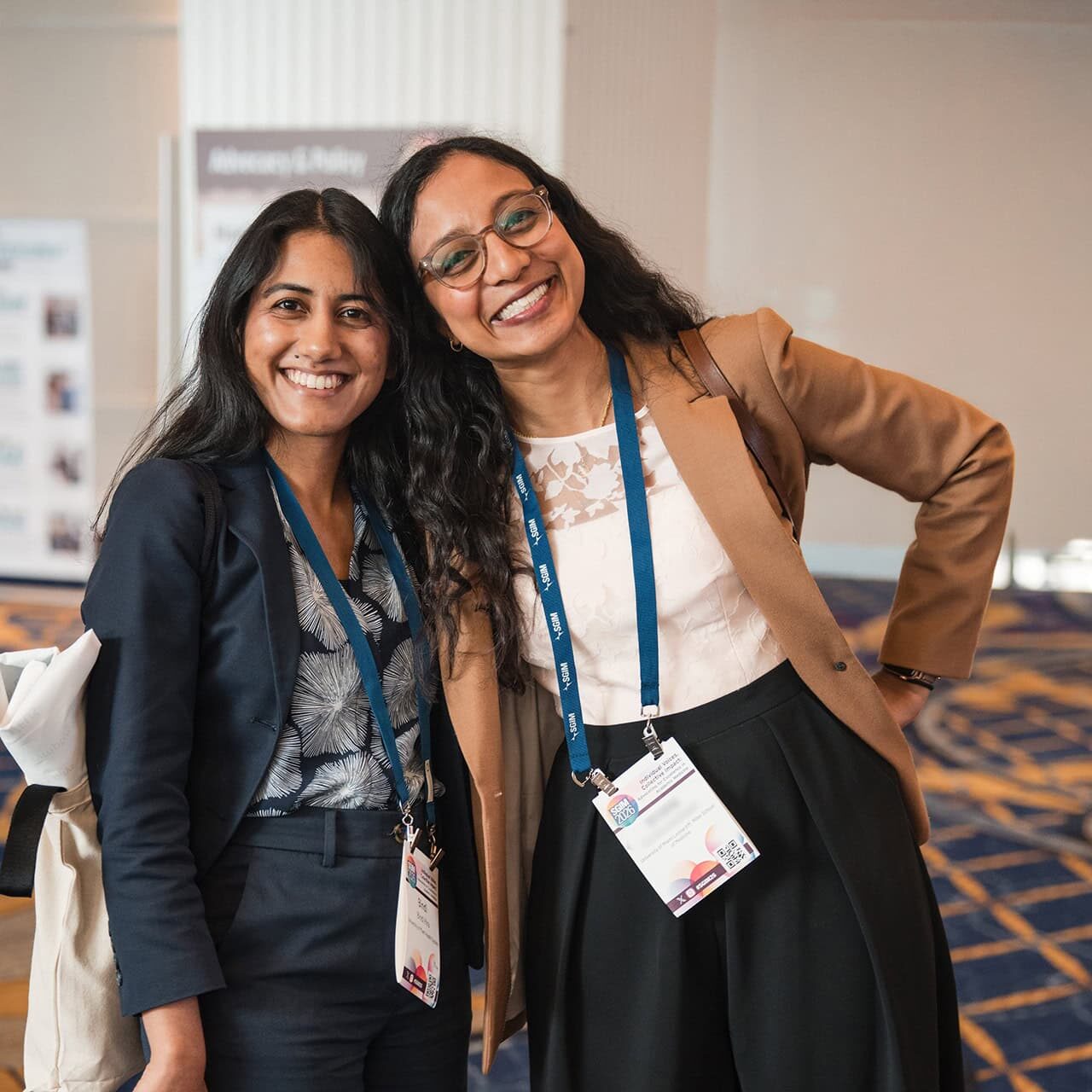 Two women smiling closely together, wearing SGIM conference badges and lanyards in a convention hallway.