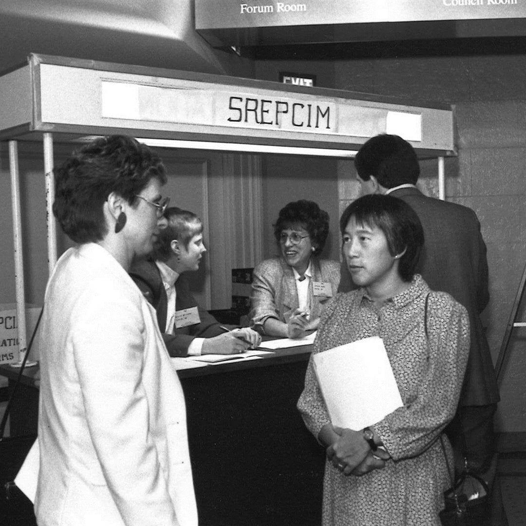 Black and white photo of two individuals engaged in conversation in front of a booth at SGIM SREPCIM event.