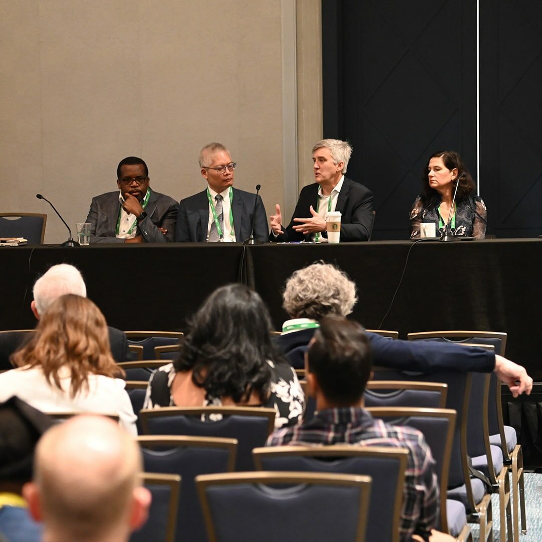 A panel of four individuals seated at a table on stage, presenting in front of an audience at an SGIM meeting.