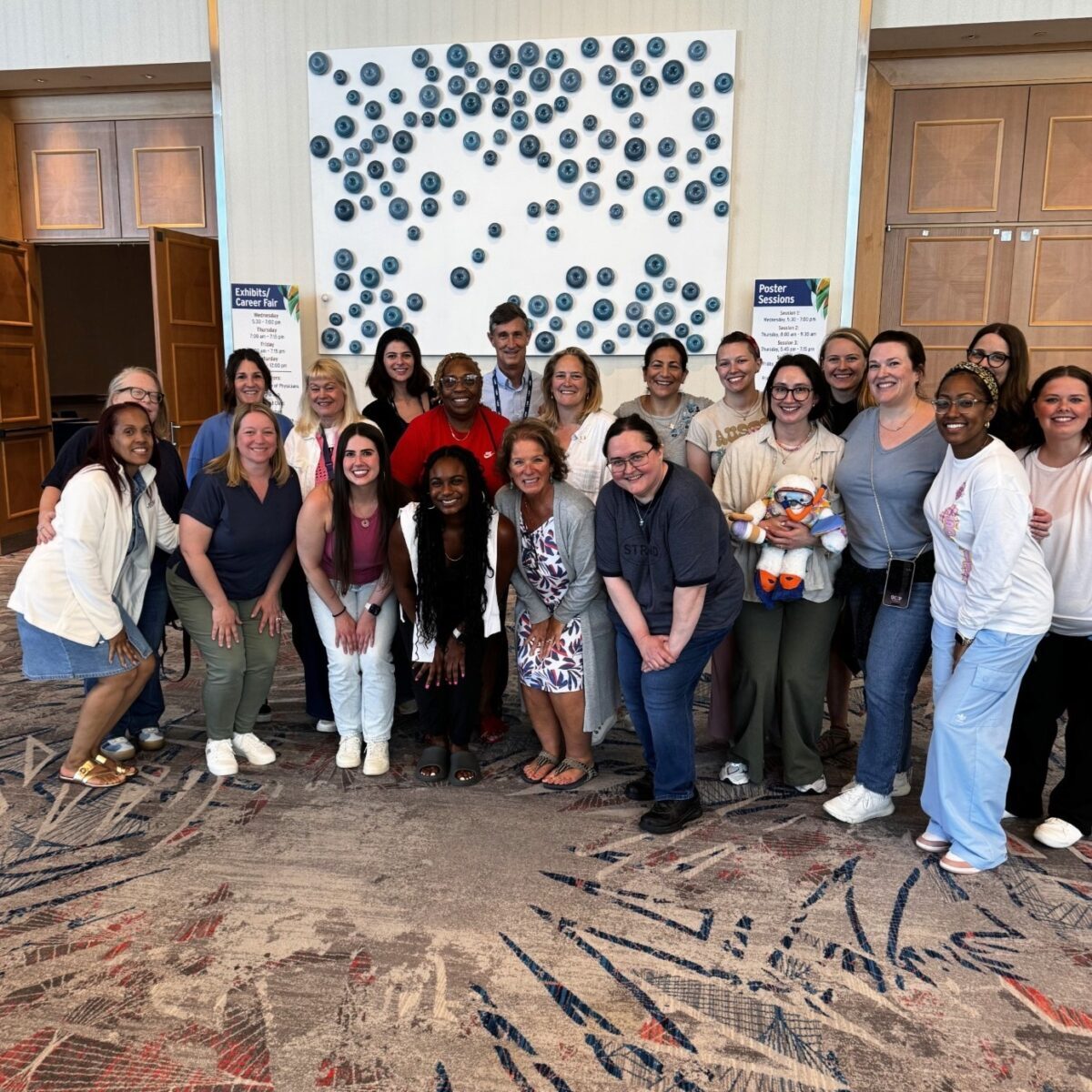 A large group of SGIM staff smiling and posing together at the SGIM25 Annual Meeting. They are standing in a hotel hallway in front of a white art installation with blue circular accents.