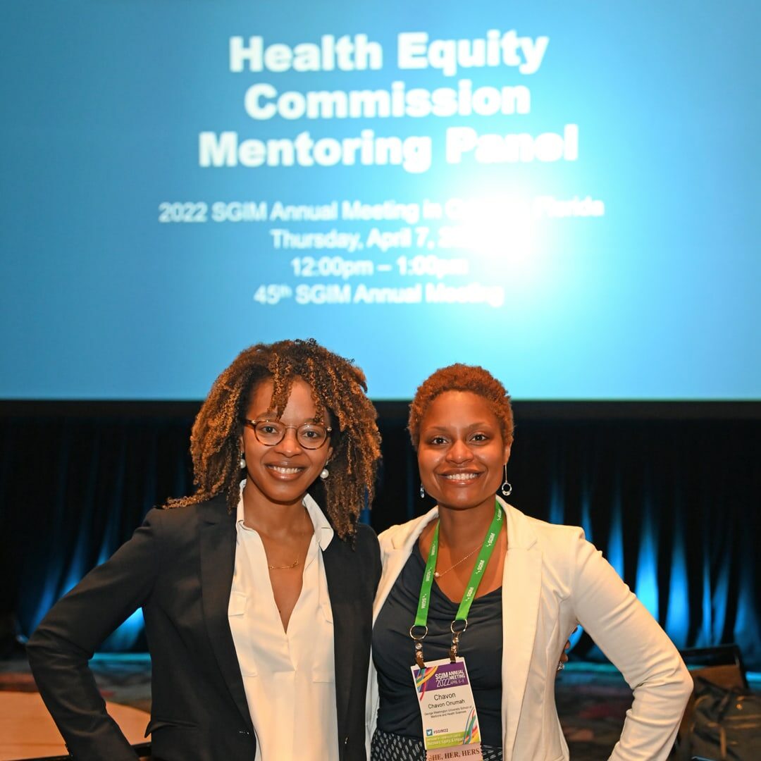 Two attendees at the 2022 SGIM Annual Meeting pose together in front of the Health Equity Commission Mentoring Panel screen.