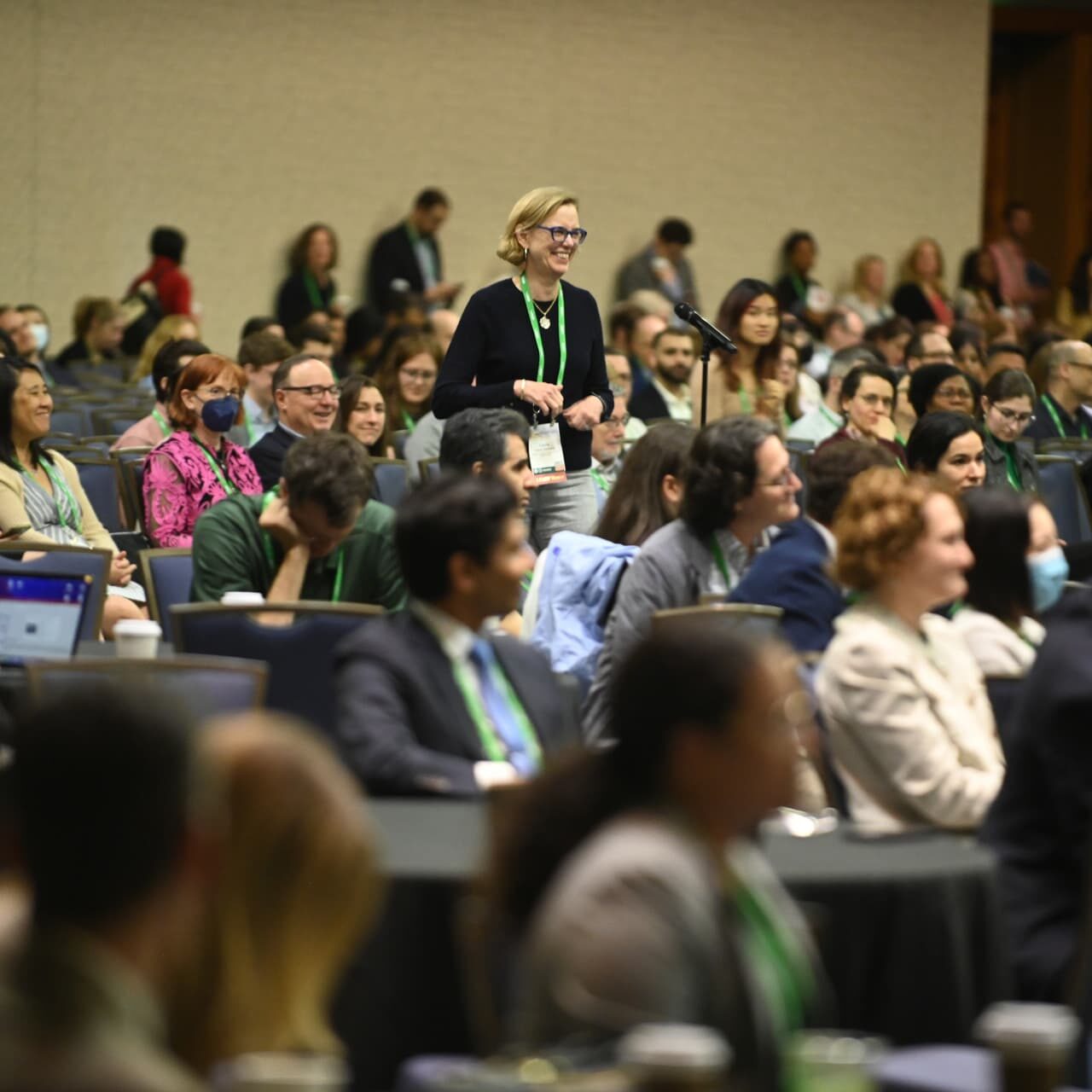 A woman standing in a crowded meeting room, surrounded by attendees who are seated and focused on the event.