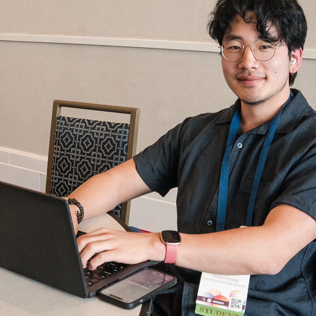 Young man in glasses and a dark shirt smiling while typing on a laptop at a conference, wearing a blue lanyard and student badge.