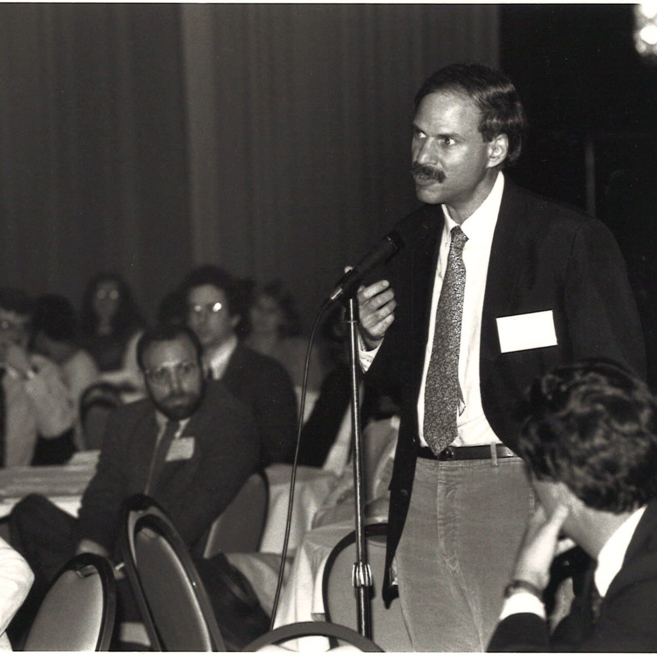 An early black and white image of a SREPCIM meeting, featuring an attendee standing at a microphone and addressing the room. SREPCIM was the original name of the Society of General Internal Medicine.