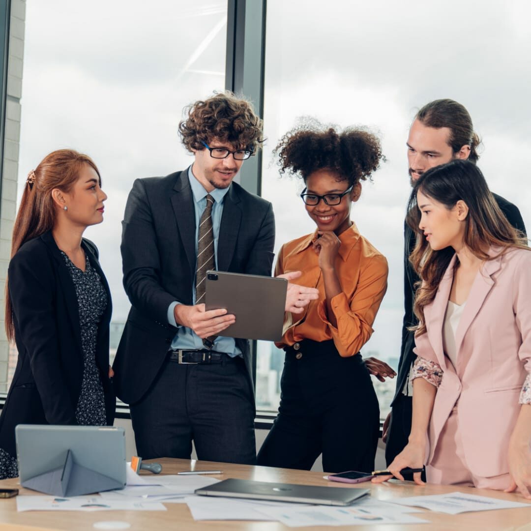 Stock image featuring five corporate professionals engaged in a meeting, viewing content on a tablet.