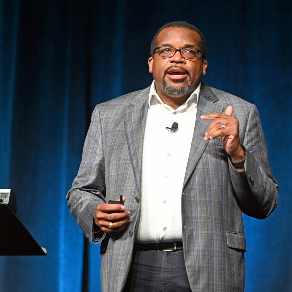 An individual wearing a suit and glasses, engaged in public speaking as an event speaker.