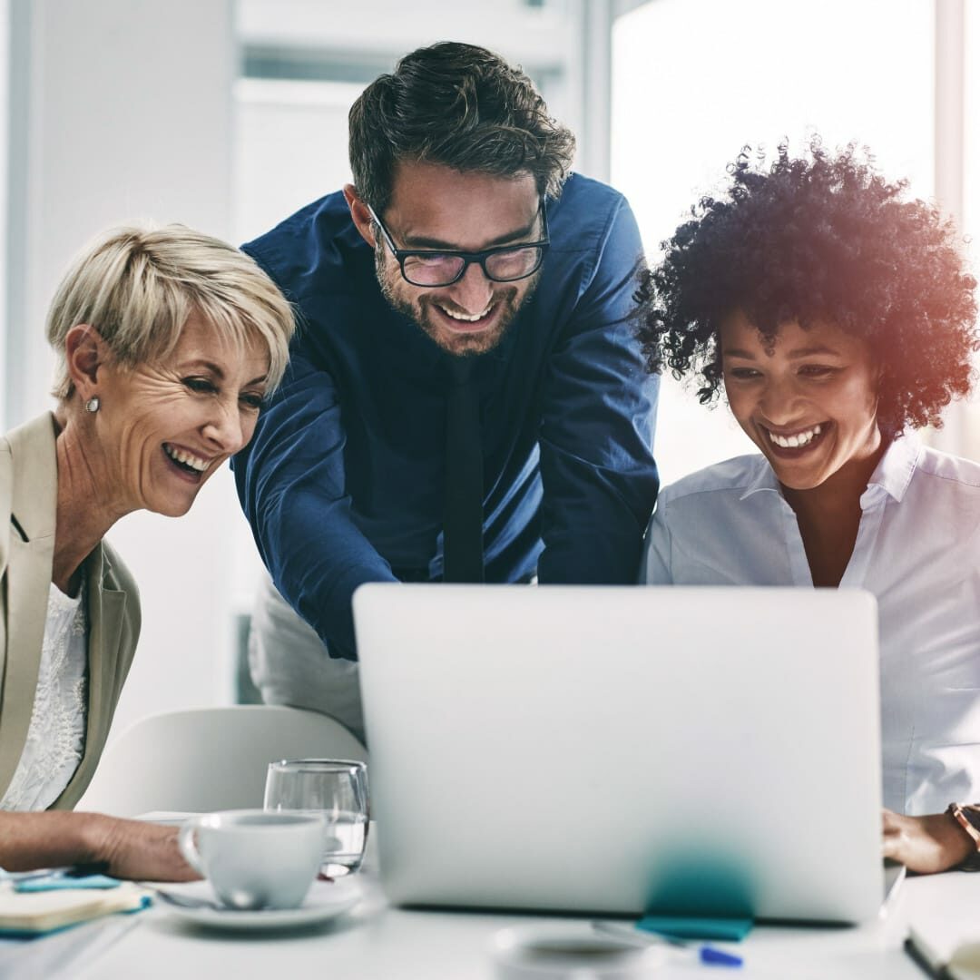 Stock image of three smiling individuals looking at a laptop in a corporate office.